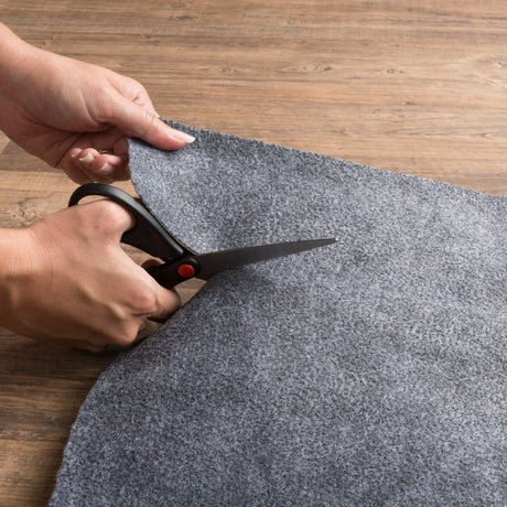 Gray rug pad edge trimming with scissors, hands adjusting the underlay size
