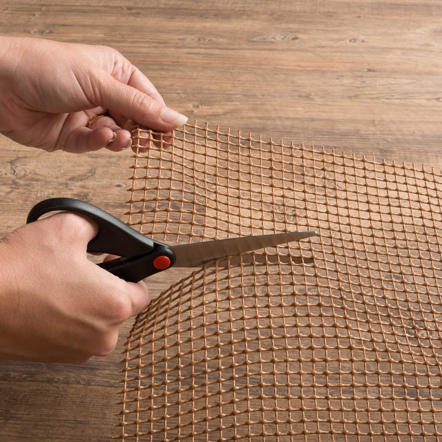 Outdoor rug pad with tan square mesh grid being trimmed with scissors